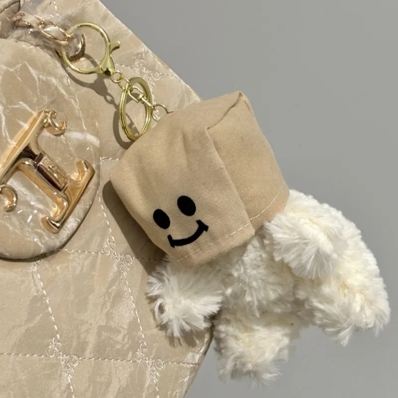 Plush dog keychain with paper bag mask featuring smiley face attached to beige quilted handbag