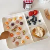 White divided plate with miniature pastries, blueberries, cherry tomatoes, and bread roll with wooden utensils